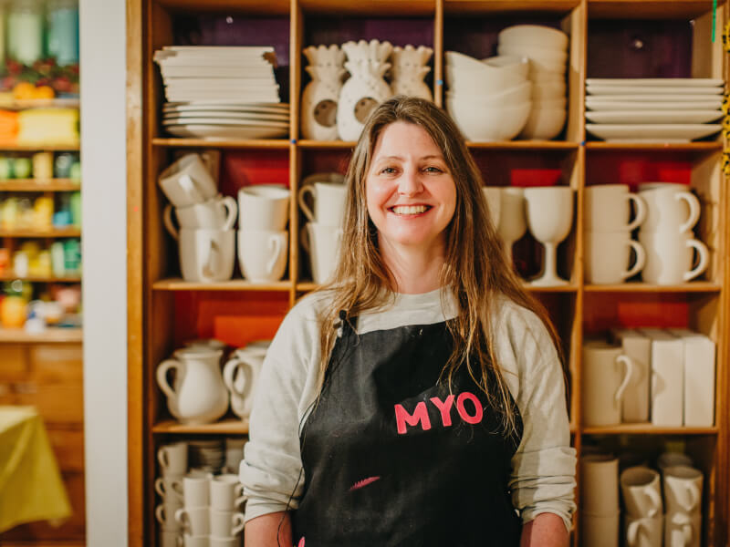 A woman wearing an apron smiling in front of shelves of pottery.